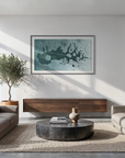 A large framed abstract art print with teal, blue, and black splatter-like brushstrokes hangs on a white wall in a modern living room setting with two gray sofas, a dark wood console, and a round black marble coffee table.