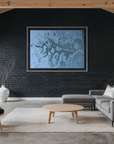 A modern living room with a large framed abstract blue and gray art print on a black brick wall, styled with a gray sectional sofa, a wooden armchair, a round coffee table, and a white vase with dried branches.