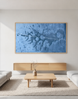 A large framed abstract blue painting hangs on a white wall in a modern living room with two light gray sofas, a wooden coffee table, and a woven rug.