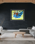 A framed abstract painting with blue, yellow, orange, and black brushstrokes hangs on a black brick wall in a modern loft-style living room with a gray sectional sofa, wooden chair, and exposed wood beams.