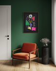 A framed abstract art print with colorful geometric shapes hangs on a dark green wall above a rust-orange armchair, next to a black marble side table with a vase of flowers.