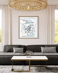 A framed abstract art print with blue and black lines on a white background, hanging on a beige wall in a modern living room with a dark gray sectional sofa, gold-framed coffee table, and large windows.