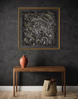A framed abstract art print with white, gold, and black scribbled lines on a dark background, hanging on a dark textured wall above a wooden console table with a terracotta vase and a woven basket.