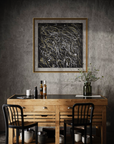 A framed abstract black-and-white artwork with gold lines hangs on a textured gray wall above a wooden console table with two black chairs, bottles, and a vase in a moody interior setting.