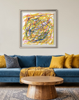A framed abstract art print with colorful scribbled lines in yellow, blue, red, and orange on a white background, hanging on a beige wall above a blue velvet sofa with yellow pillows in a styled living room.