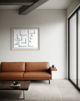 A modern living room scene featuring a mid-century style tan leather sofa with a side table, a dark wood coffee table, and a framed abstract geometric print on a textured off-white wall.