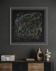 A framed abstract art print featuring chaotic scribbled lines in white, yellow, and green on a black background, hung on a dark gray textured wall above a wooden console table with decorative objects.