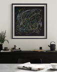 A framed abstract art print with chaotic white, yellow, and green scribbled lines on a black background, hanging on a white wall above a dark wooden console table in a modern dining room setting.
