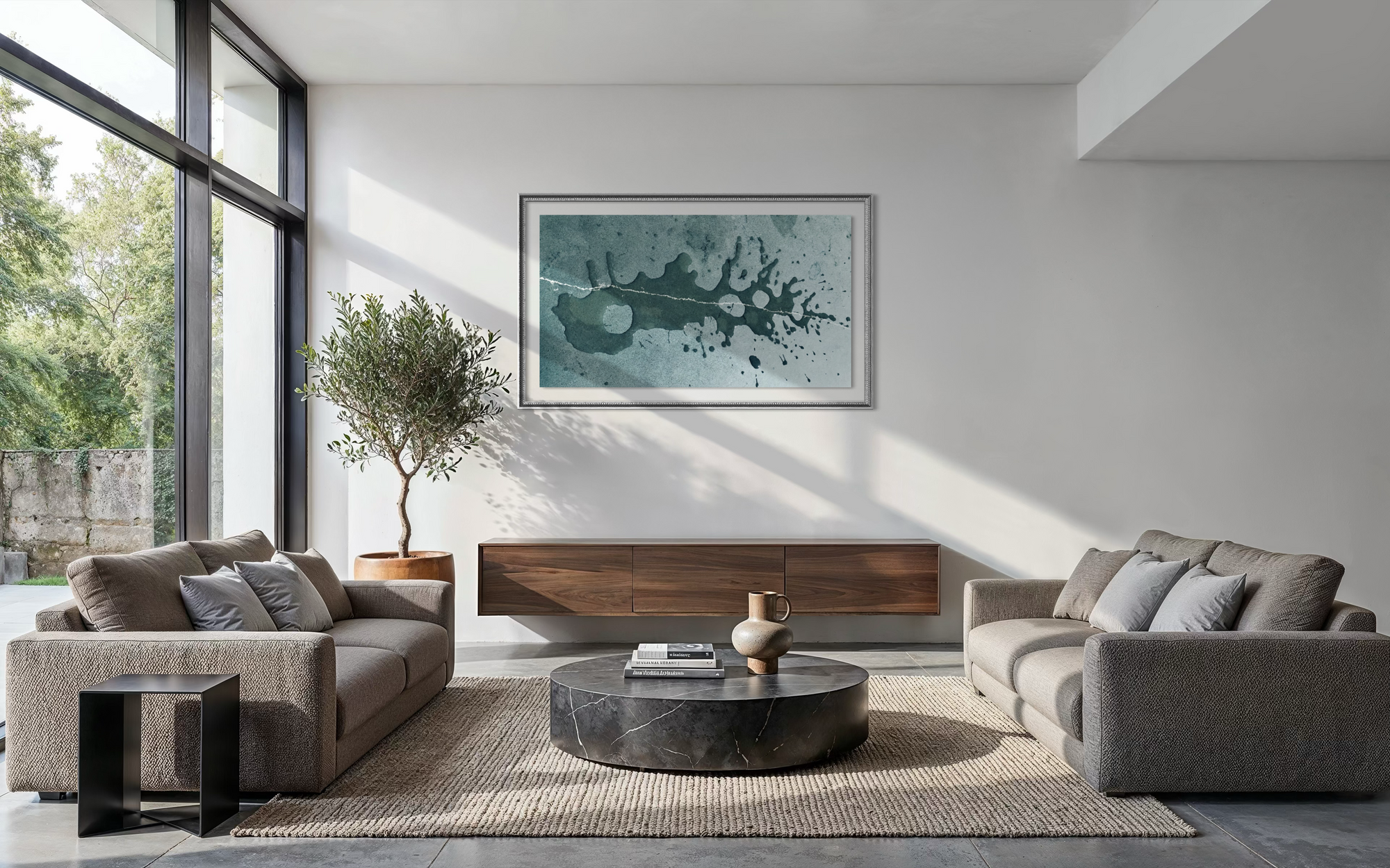 A large framed abstract art print with teal, blue, and black splatter-like brushstrokes hangs on a white wall in a modern living room setting with two gray sofas, a dark wood console, and a round black marble coffee table.