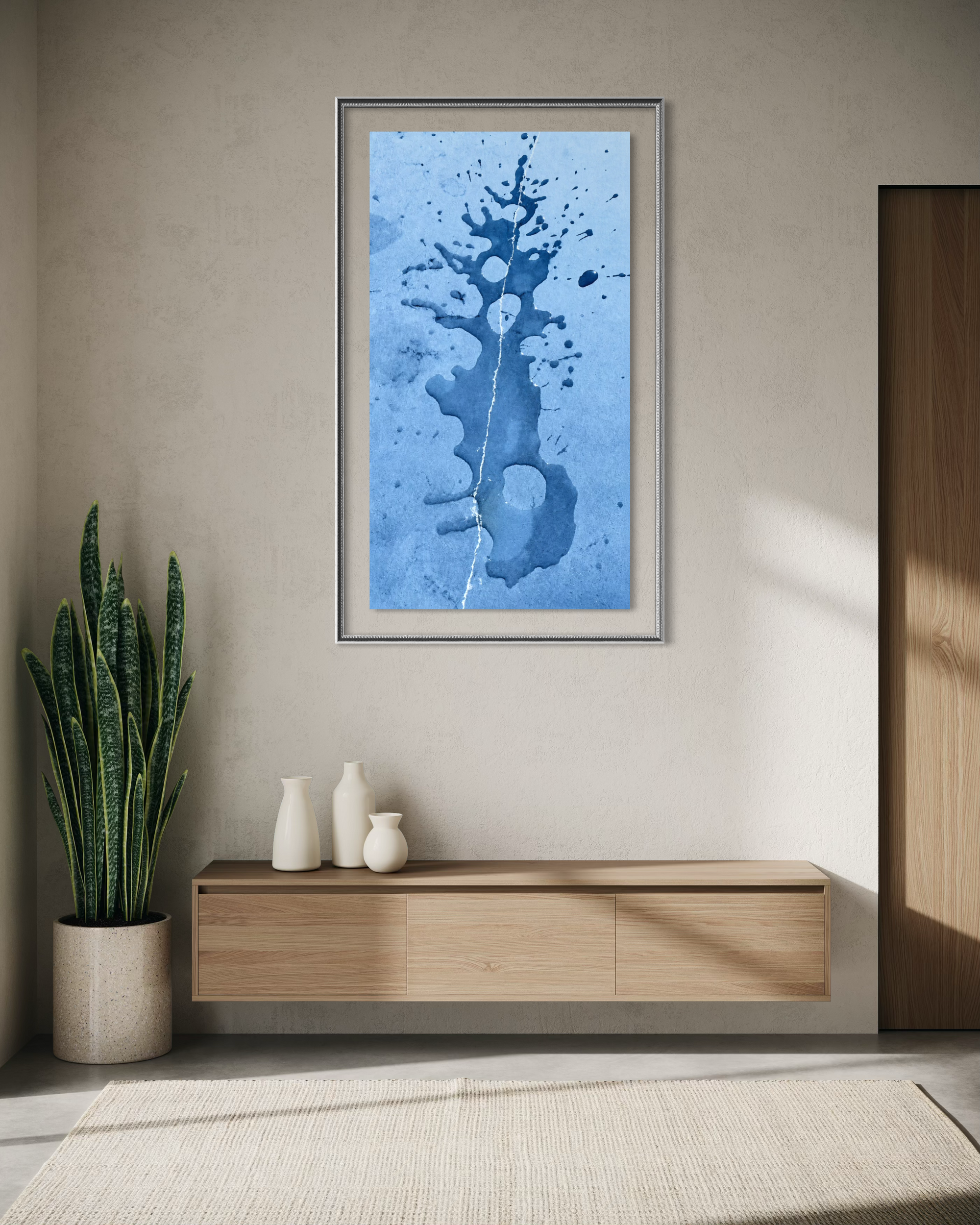 A framed vertical art print featuring an abstract blue watercolor splash design, displayed on a beige wall above a light wood console table in a modern, sunlit room.