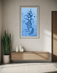 A framed vertical art print featuring an abstract blue watercolor splash design, displayed on a beige wall above a light wood console table in a modern, sunlit room.