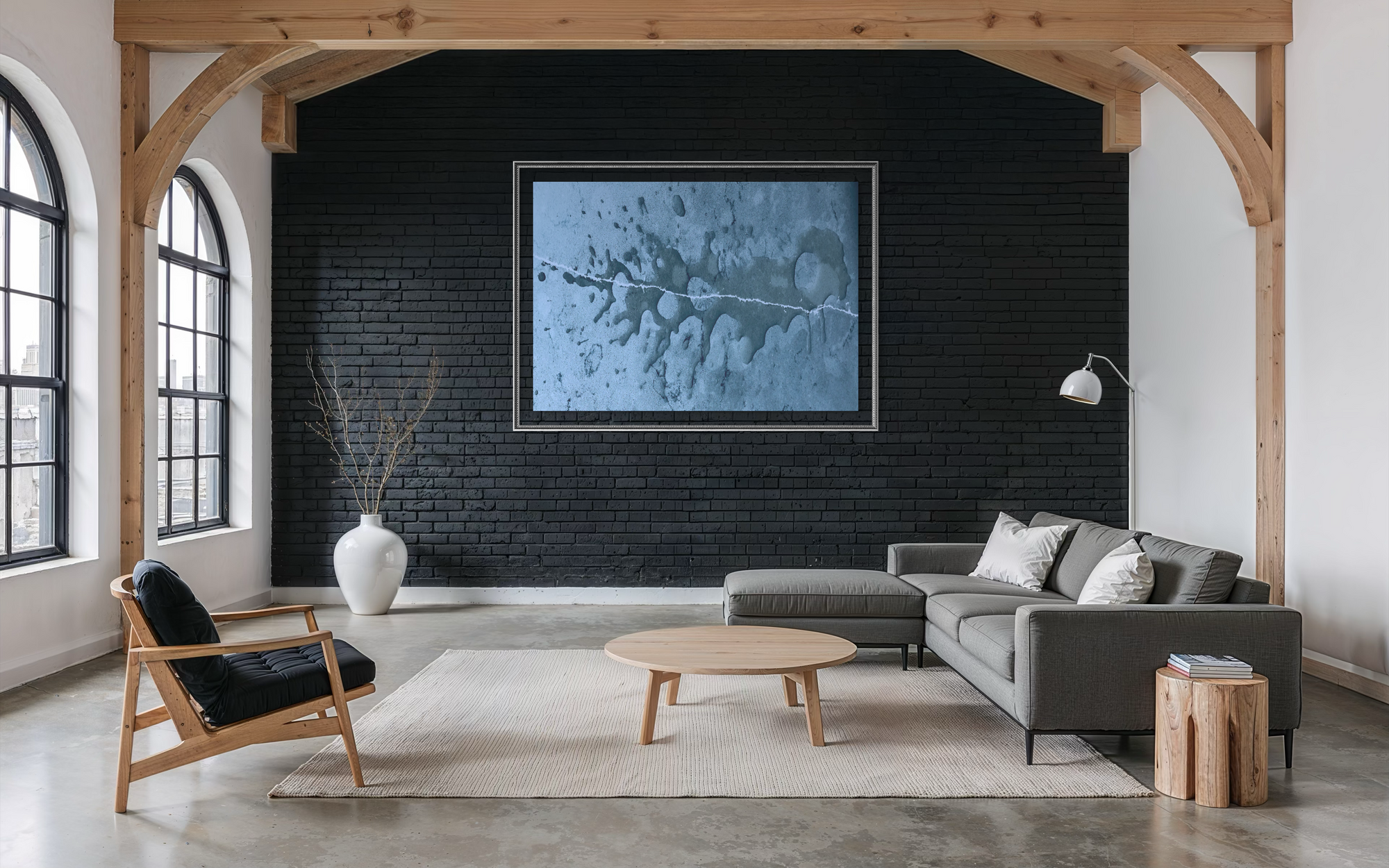 A modern living room with a large framed abstract blue and gray art print on a black brick wall, styled with a gray sectional sofa, a wooden armchair, a round coffee table, and a white vase with dried branches.
