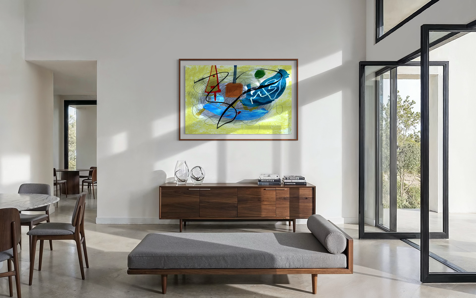 A framed abstract painting with blue, yellow, orange, and black brushstrokes hangs on a white wall in a modern dining room with a gray settee, chairs set up around a table with open glass doors and abundant light