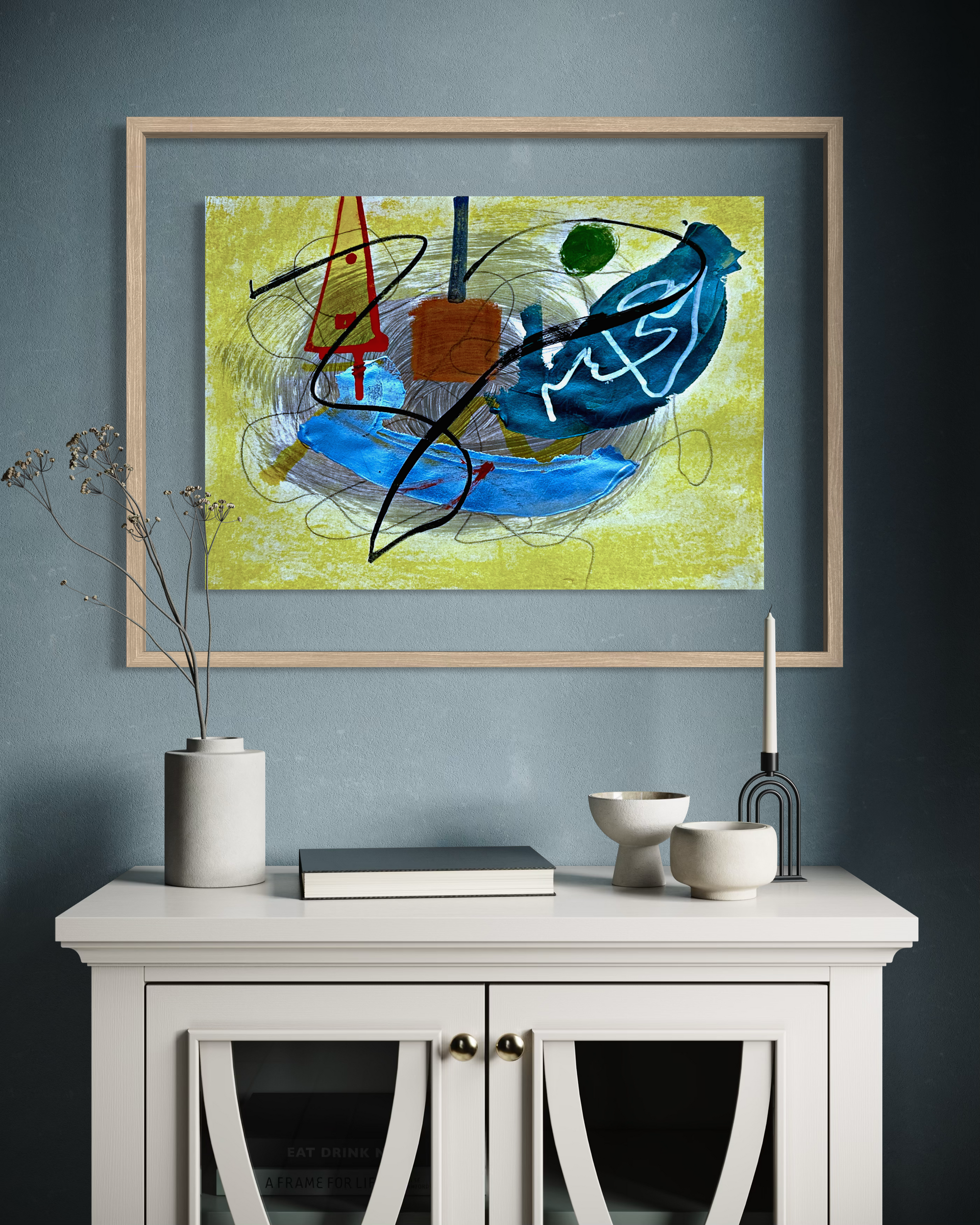 An abstract framed painting featuring blue, yellow, black, and red brushstrokes, displayed on a dark blue-gray wall above a white sideboard with decorative ceramic objects.
