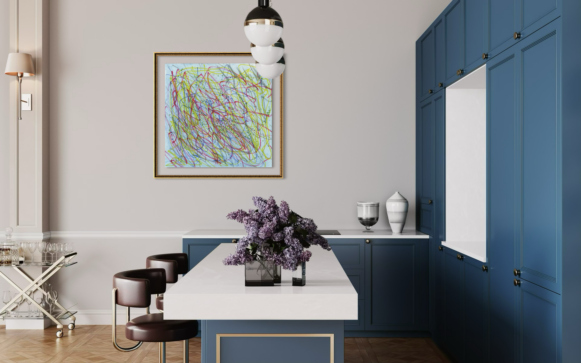 A framed abstract art print with multicolored scribbled lines hangs on a light gray wall in a modern kitchen setting with blue cabinets, a white island, and a vase of purple flowers in the foreground.