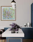 A framed abstract art print with multicolored scribbled lines hangs on a light gray wall in a modern kitchen setting with blue cabinets, a white island, and a vase of purple flowers in the foreground.