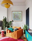 A cozy living room corner featuring a bright orange armchair with a teal throw blanket, a colorful abstract line art print on the wall, a large yellow pendant light, and potted plants.