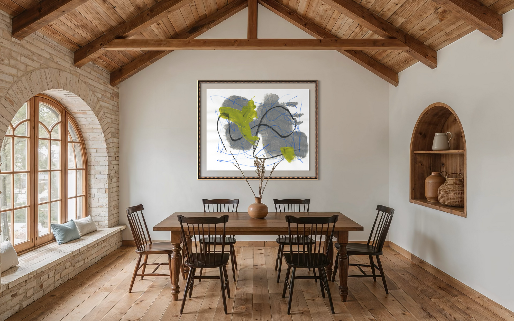 A dining room interior with a wooden dining table and six black spindle-back chairs, set under a high wood-beamed ceiling. A framed abstract print hangs on the back wall above the table, and a built-in arched niche on the right holds ceramic vases.