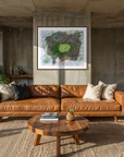 A modern living room featuring a tan leather sofa with decorative throw pillows, set on a woven jute rug in front of a concrete wall with a framed abstract painting. A round wooden coffee table sits in the foreground, flanked by potted plants, and a dining area is visible in the background.