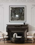 A framed abstract black and white artwork hangs above a dark fireplace in a styled living room with two white armchairs, a round dark coffee table, and a potted plant against ornate white wall molding.