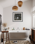 A modern bathroom interior featuring a freestanding oval bathtub centered beneath a framed abstract print on a white wall. Two woven pendant lights hang above, with a wooden stool, patterned rug, toiletries, and a window visible in the room.