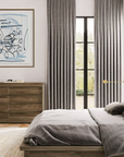 A styled bedroom interior featuring a bed with gray bedding and a light wood frame, matching gray curtains, a wooden dresser, a white armchair, and a blue abstract wall art print.
