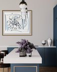 A framed abstract art print with blue and black scribbles on a white background, hanging on a light gray wall in a modern kitchen with blue cabinets and a white island.