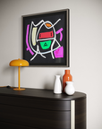 A framed abstract geometric wall art print hangs on a light gray wall above a dark wood console table. The artwork features bold, colorful shapes on a black background, including pink, green, orange, red, and white.
