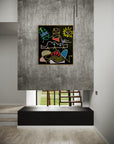 A framed abstract art print with colorful geometric shapes on a black background, displayed on a textured gray wall in a modern interior setting.