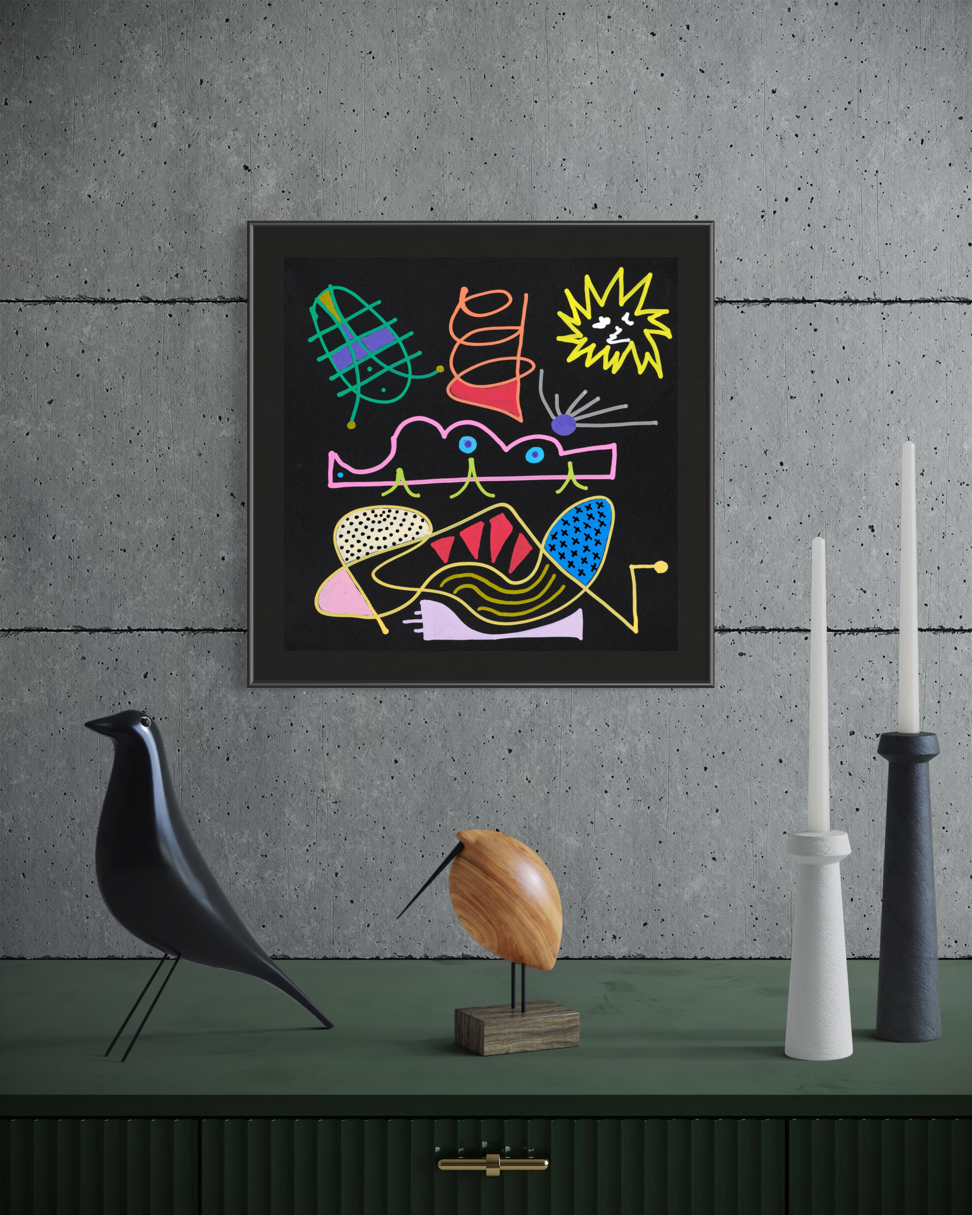 A framed abstract artwork with colorful geometric and organic shapes on a black background, displayed on a dark green console against a textured gray wall. Decorative objects including a black bird sculpture, a wooden bird figurine, and white and black candlesticks sit below.