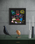 A framed abstract artwork with colorful geometric and organic shapes on a black background, displayed on a dark green console against a textured gray wall. Decorative objects including a black bird sculpture, a wooden bird figurine, and white and black candlesticks sit below.