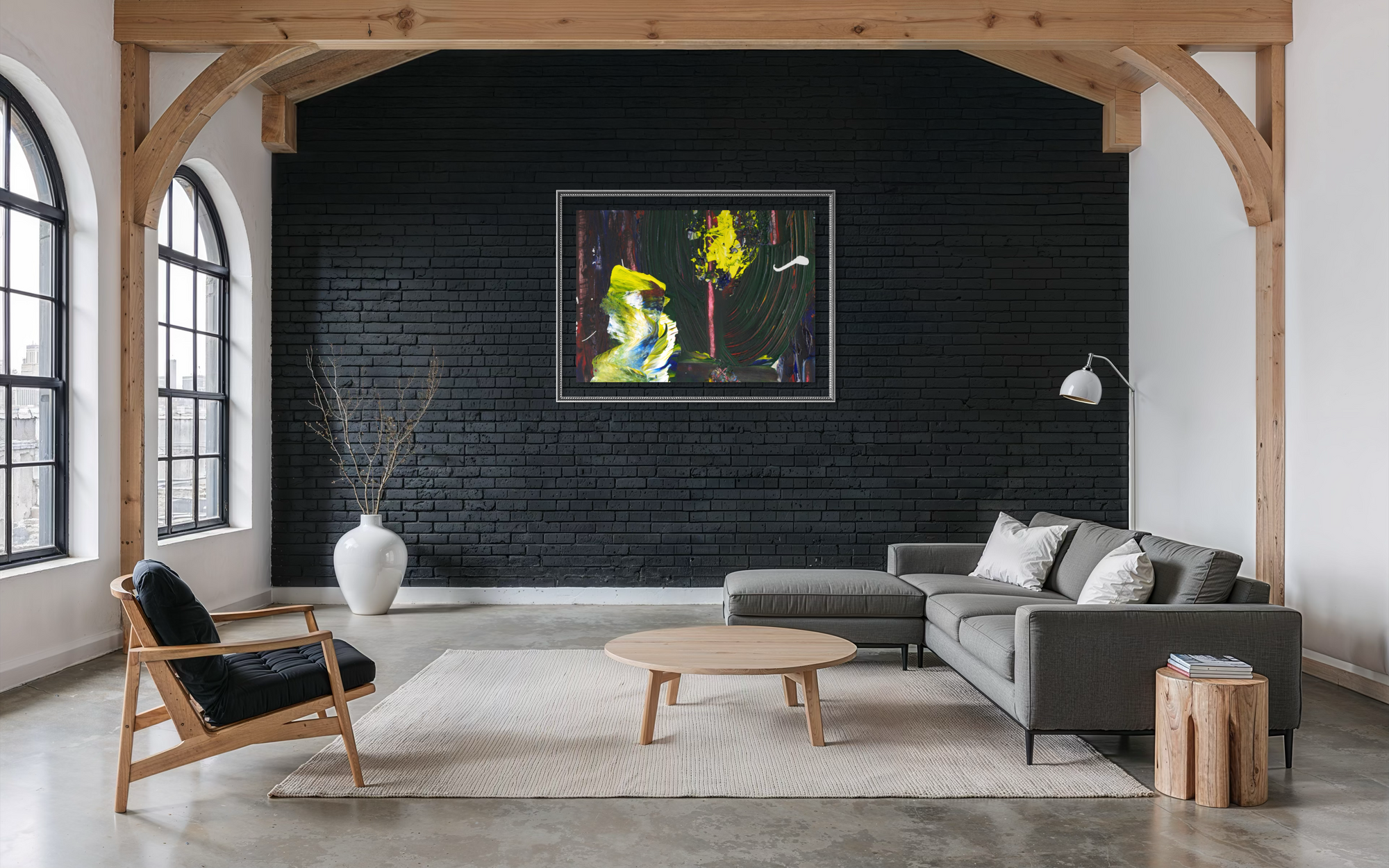 A framed abstract painting with vibrant yellow, green, and blue brushstrokes hangs on a black brick wall in a modern living room with a gray sectional sofa, wooden chair, and exposed wood beams.