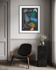 A framed abstract art print with colorful organic shapes on a dark background, displayed on a gray wall in a modern room setting above a black chair and next to a black marble side table with a vase of flowers.