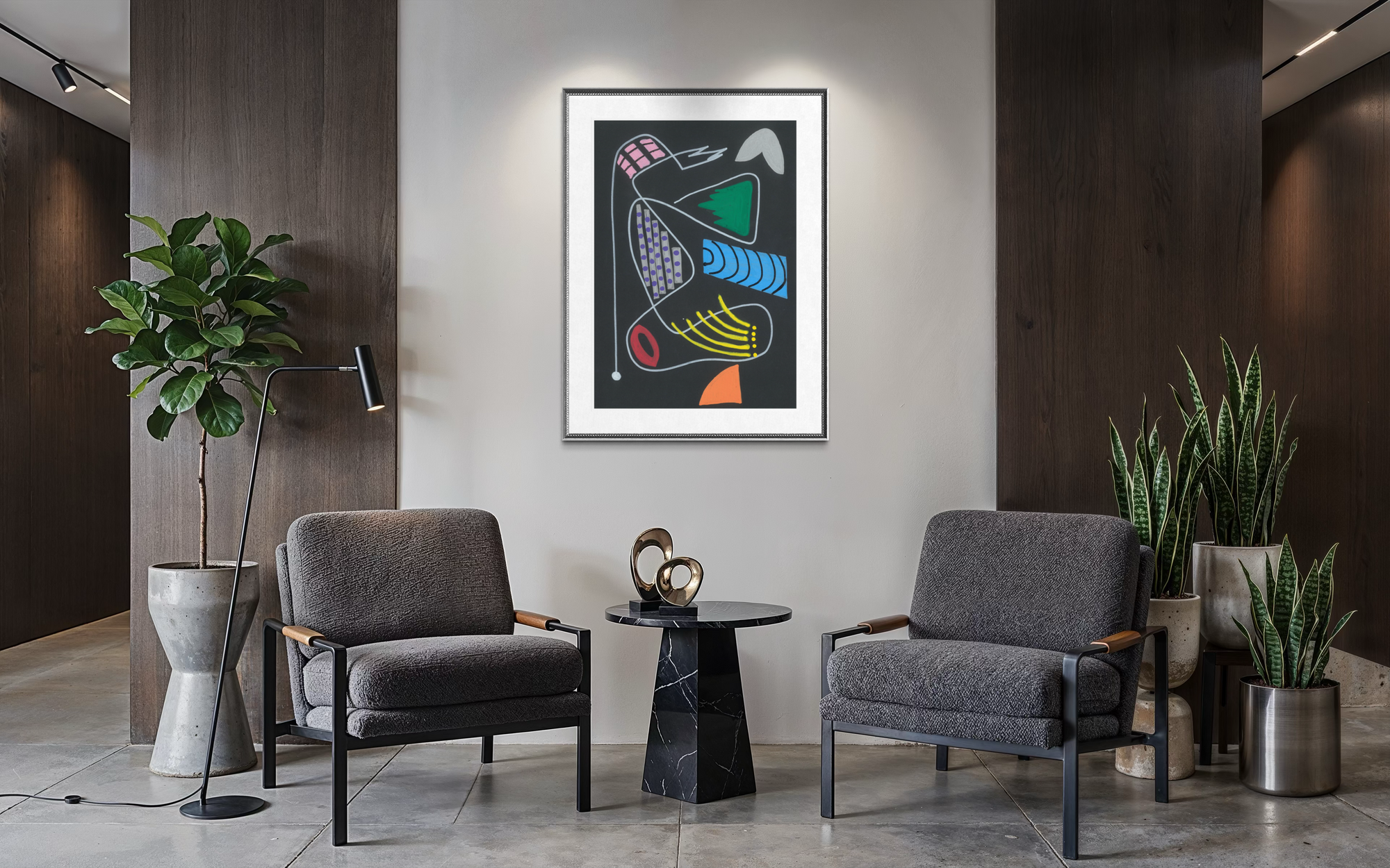 A framed abstract art print with colorful geometric shapes on a dark background hangs on a light gray wall in a modern living room setting with two gray armchairs, a small black marble side table, and potted plants.