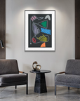 A framed abstract art print with colorful geometric shapes on a dark background hangs on a light gray wall in a modern living room setting with two gray armchairs, a small black marble side table, and potted plants.