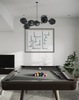 A modern pool table in a minimalist room setting, featuring a dark wood frame, gray felt surface, a rack of colorful billiard balls, a cue stick, and a white cue ball. Above the table hangs a black multi-globe chandelier, and a framed abstract print is mounted on the white wall behind it.