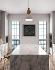 A modern kitchen interior with a large white marble island in the foreground, white cabinetry, wood floors, and a black framed abstract artwork centered between two tall windows.